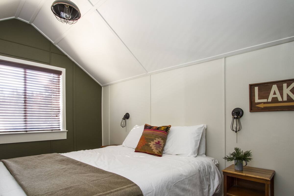 Une chambre avec un lit, une fenêtre avec des stores, une enseigne fixée au mur sur laquelle on peut lire « LAC », une table de chevet avec une plante et un luminaire fixé au mur.
