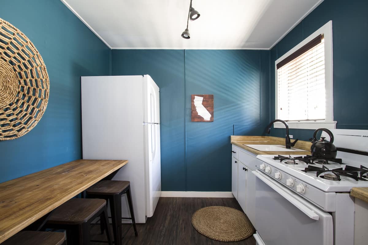 Une cuisine avec une table en bois, des tabourets de bar, un réfrigérateur blanc, une cuisinière et une fenêtre avec des stores.