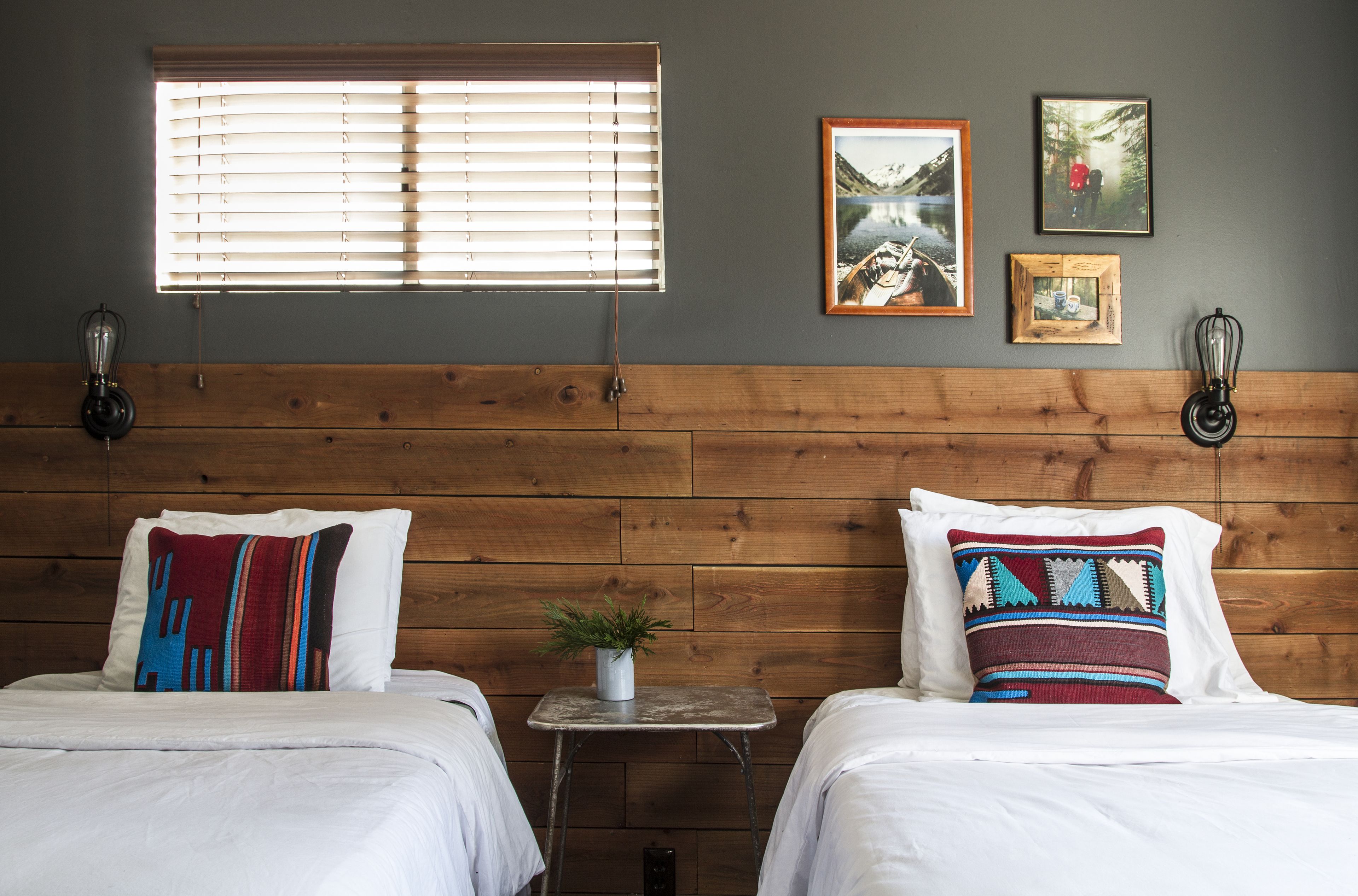 A room with two beds, a small table with a plant, and a window with blinds.