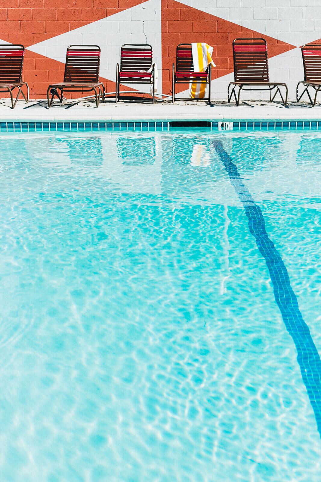 Una piscina con agua azul cristalina y un borde de azulejos. Seis sillas de metal con cojines rojos están alineadas a lo largo de la piscina.