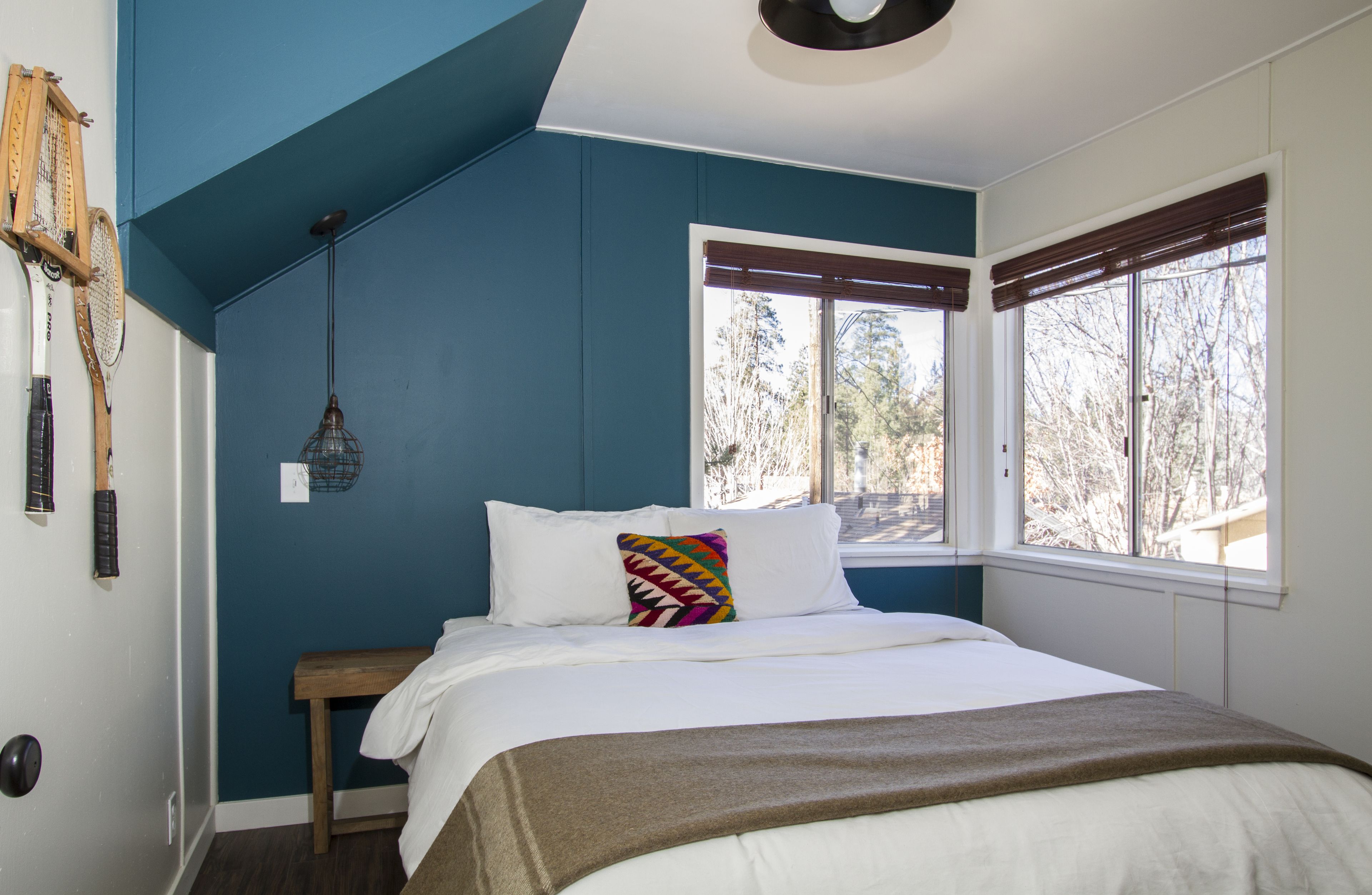 A bedroom with a sloped ceiling, a bed with white bedding, a colorful pillow, a bench, and a window with blinds.