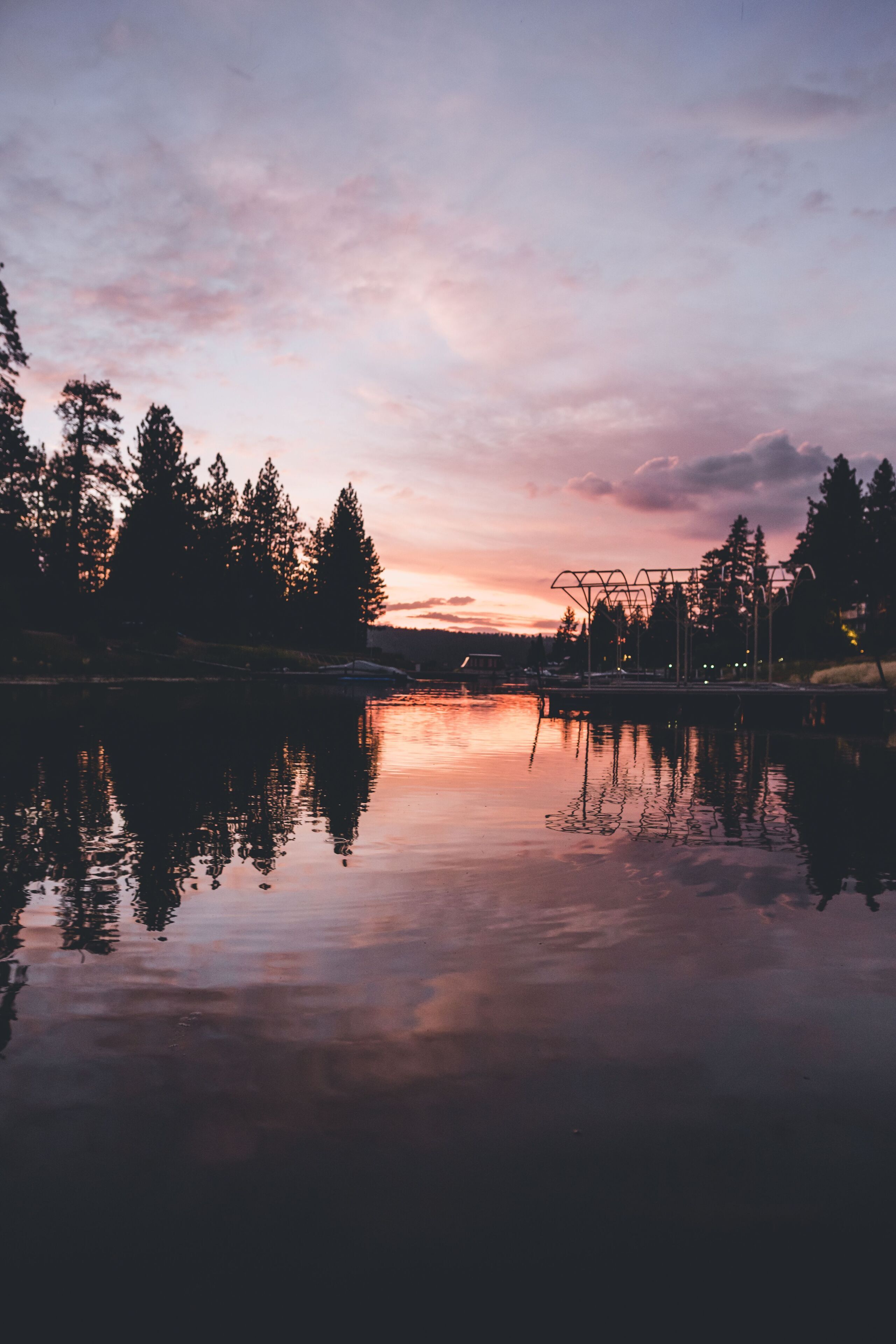 A calm lake at dusk with trees reflecting on the water, a dock, and a bridge in the background.