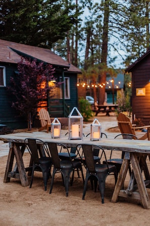 Un espacio para comer al aire libre con una mesa y sillas de madera, rodeado de árboles y una estructura que recuerda a una cabaña.