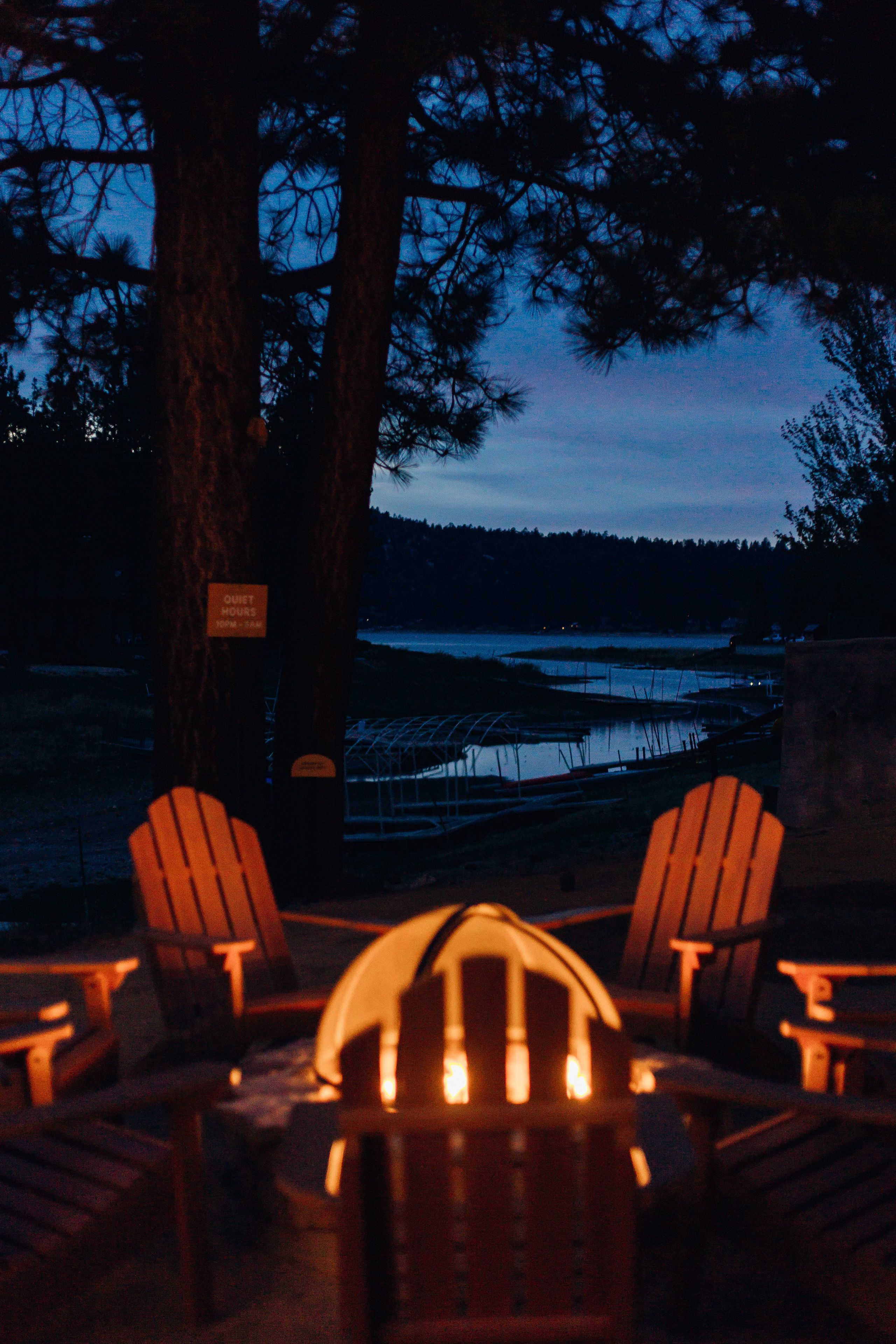 A cozy outdoor seating area with Adirondack chairs and a fire pit, overlooking a body of water and a forested area.