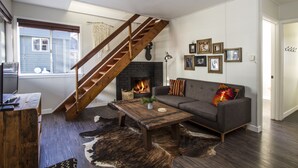 Una sala de estar con escalera de madera, chimenea, sofá gris y alfombra de cuero de vaca.