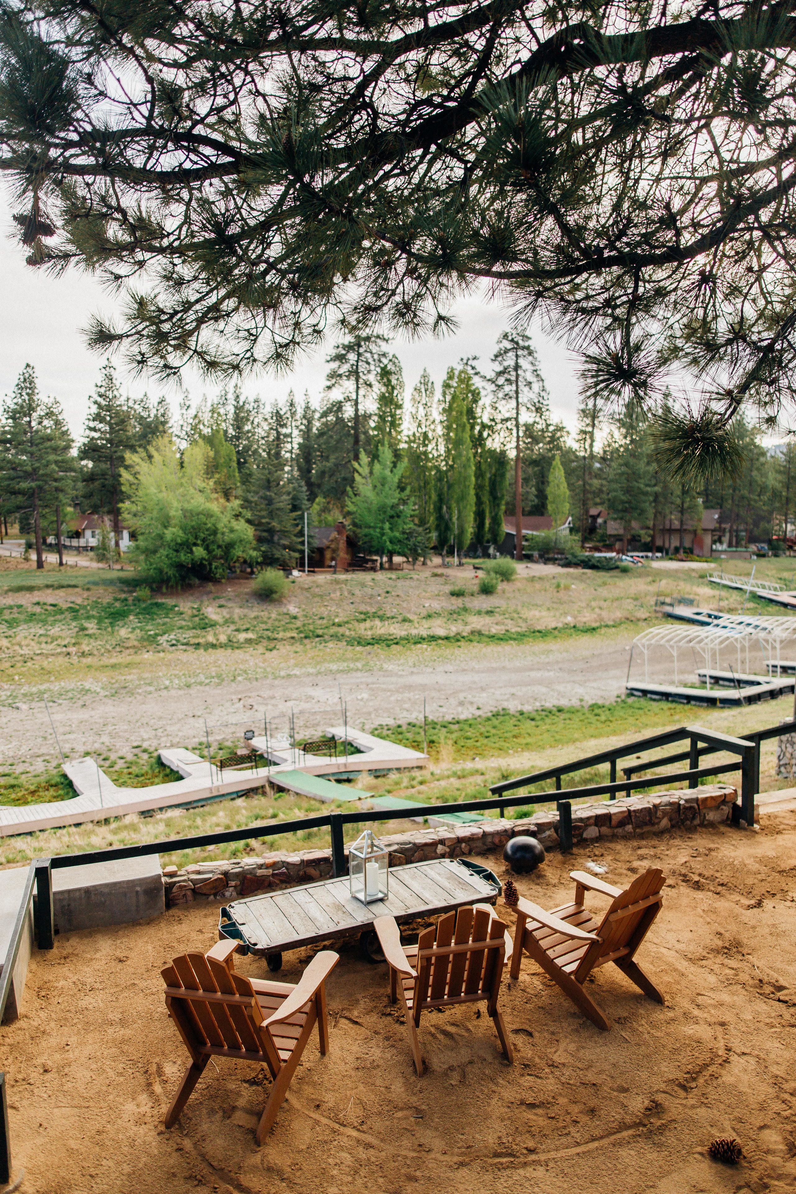 An outdoor seating area with wooden chairs and a table, overlooking a sandy landscape with trees and a dock.