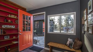 Un pasillo con una puerta roja, estantes de madera, un banco y una ventana que ofrece vistas a una casa y árboles.