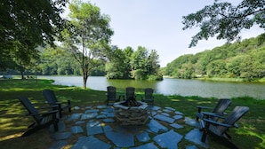 Un foyer en pierre, entouré de fauteuils Adirondack, est installé face à un lac, avec une colline boisée en arrière-plan.