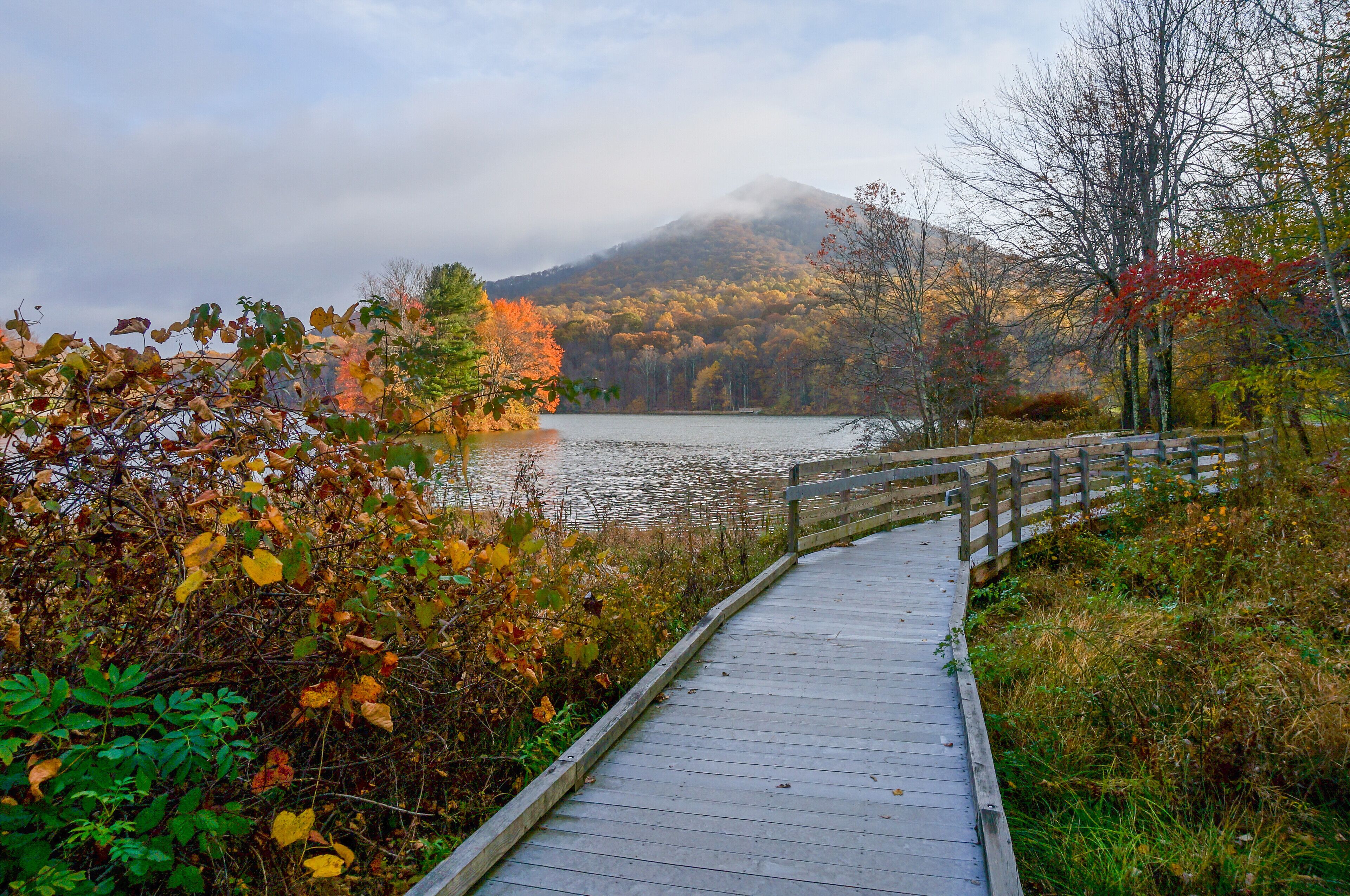 Une promenade en bois qui mène à un lac, avec des arbres aux couleurs automnales et une montagne en arrière-plan.