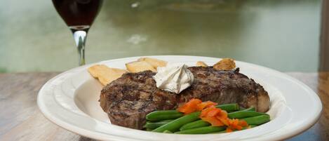 A plate of food with steak, potatoes, and green beans, and a glass of red wine on a table with a view of a body of water.