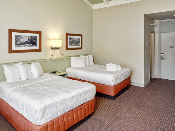 Habitación de hotel con dos camas, una mesita de noche, una lámpara y dos cuadros en la pared.