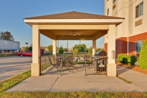 A covered outdoor seating area with tables and chairs, a barbecue grill, and a building with a brick facade in the background.