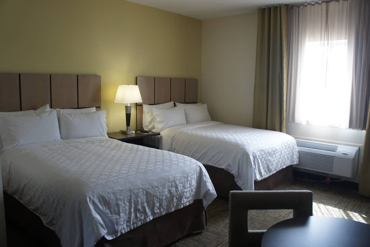 Ein Hotelzimmer mit zwei Betten, einem Nachttisch mit Lampe, einem Sessel und einem Fenster mit Vorhängen.