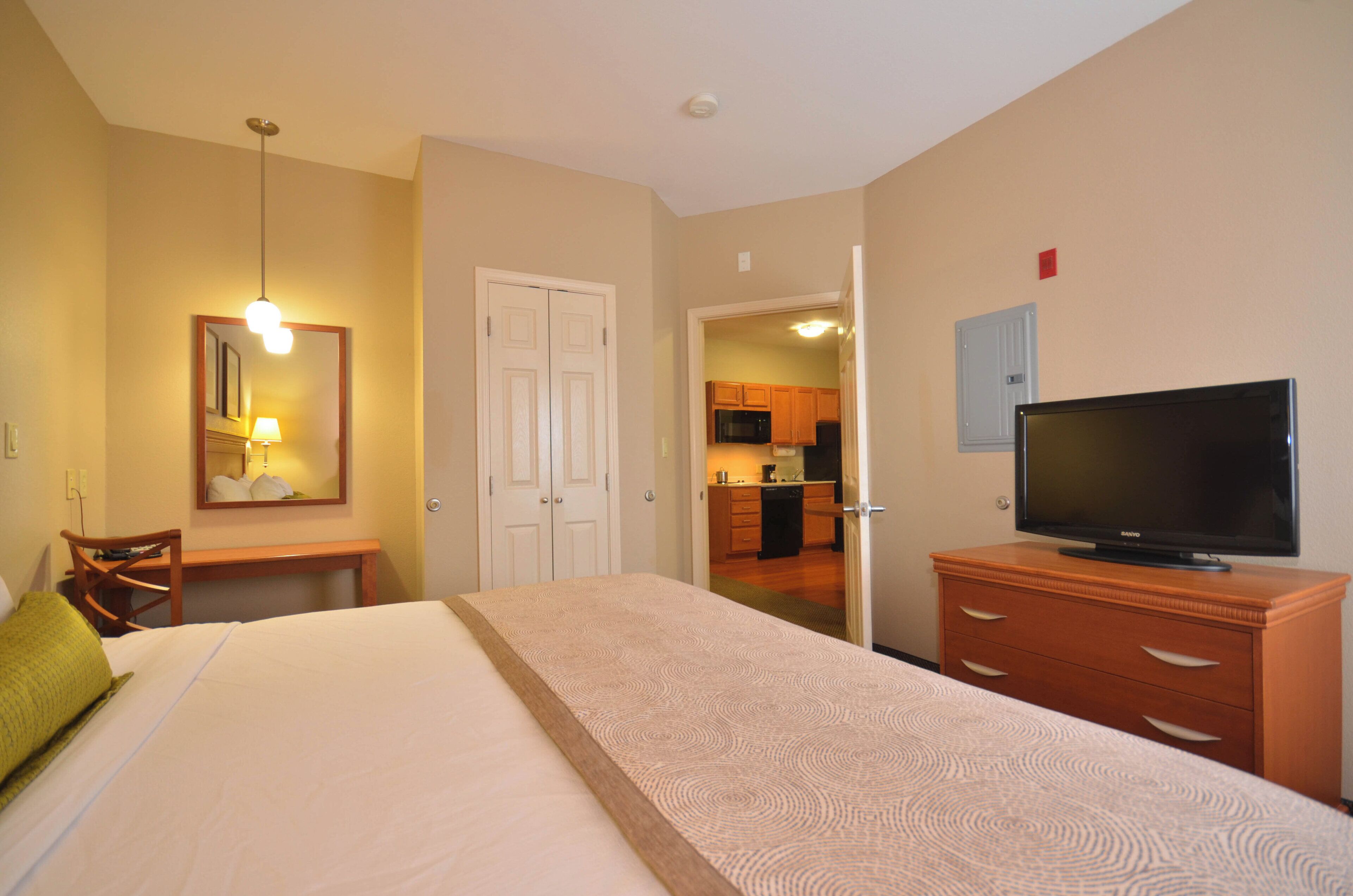 A hotel room with a bed, a television, a desk with a lamp, a chair, and a view into the kitchen.