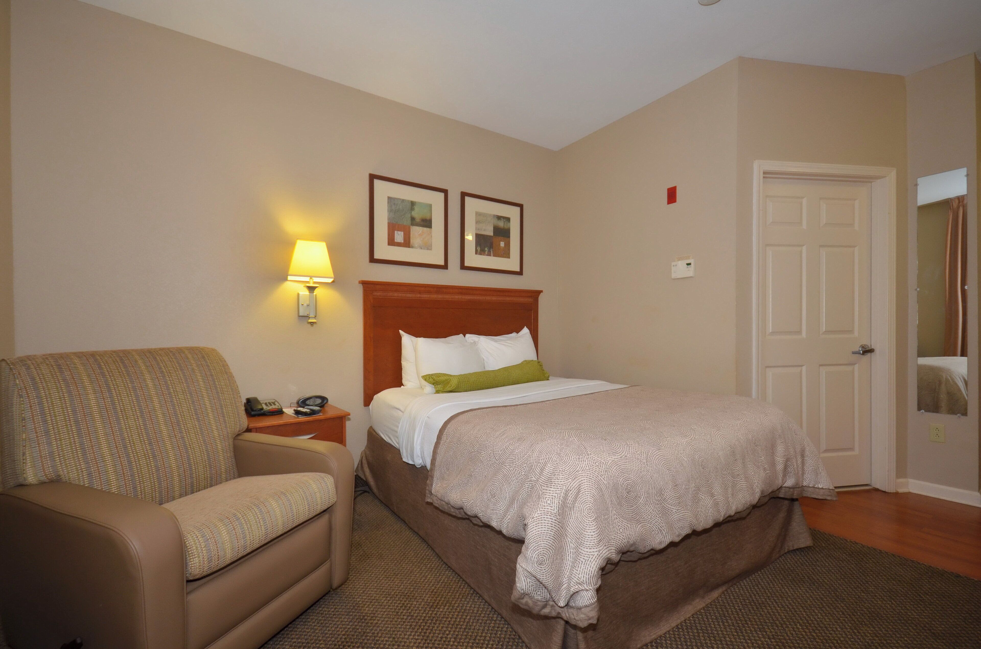 A hotel room with a bed, a chair, a nightstand with a lamp, and two framed pictures on the wall.