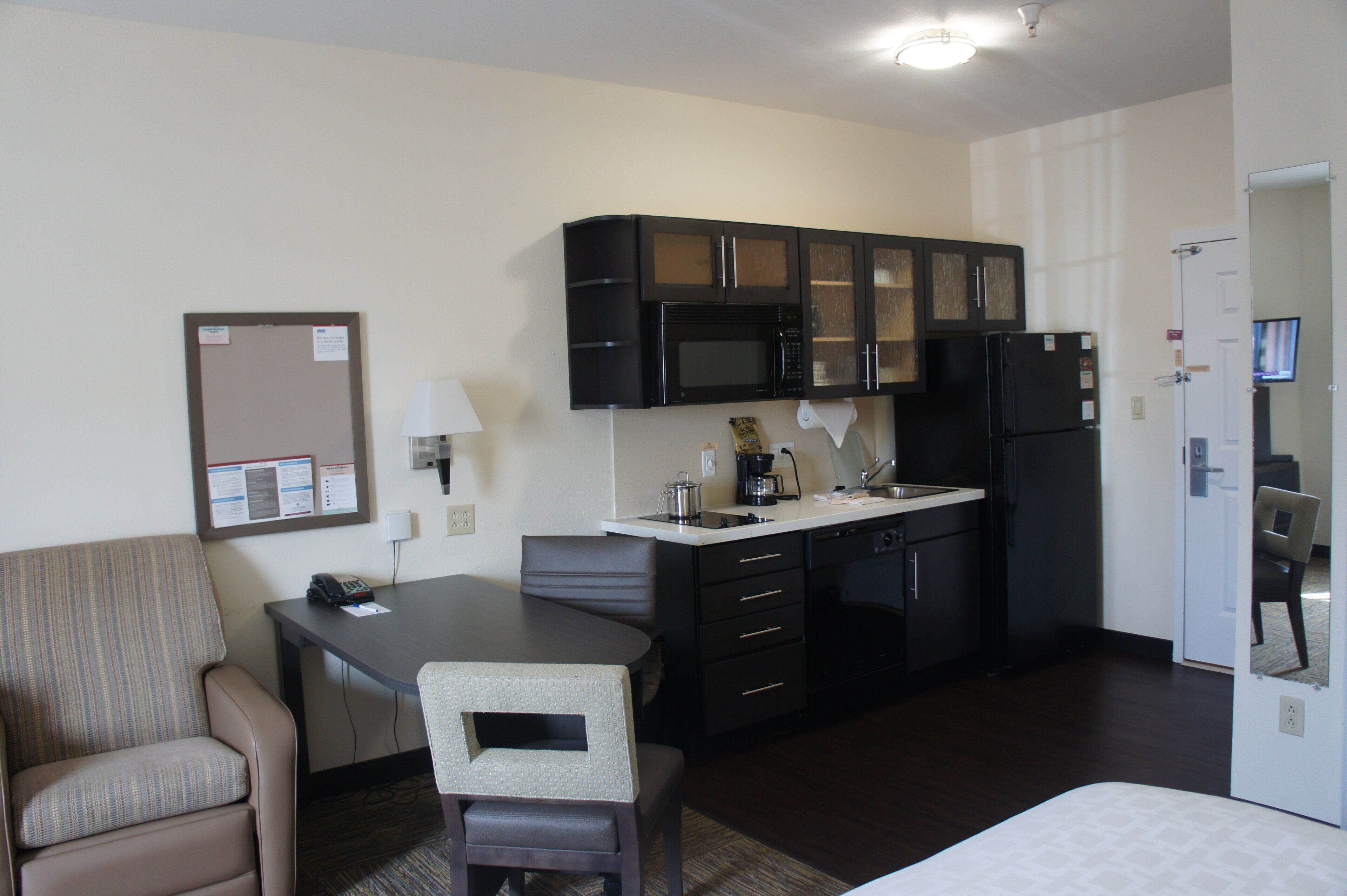 A compact kitchenette with a microwave, refrigerator, and a small sink. There is a dining area with a table and chairs, and a bulletin board on the wall.