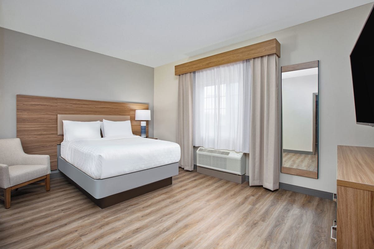 A hotel room with a bed, a chair, a mirror, and a window with curtains.