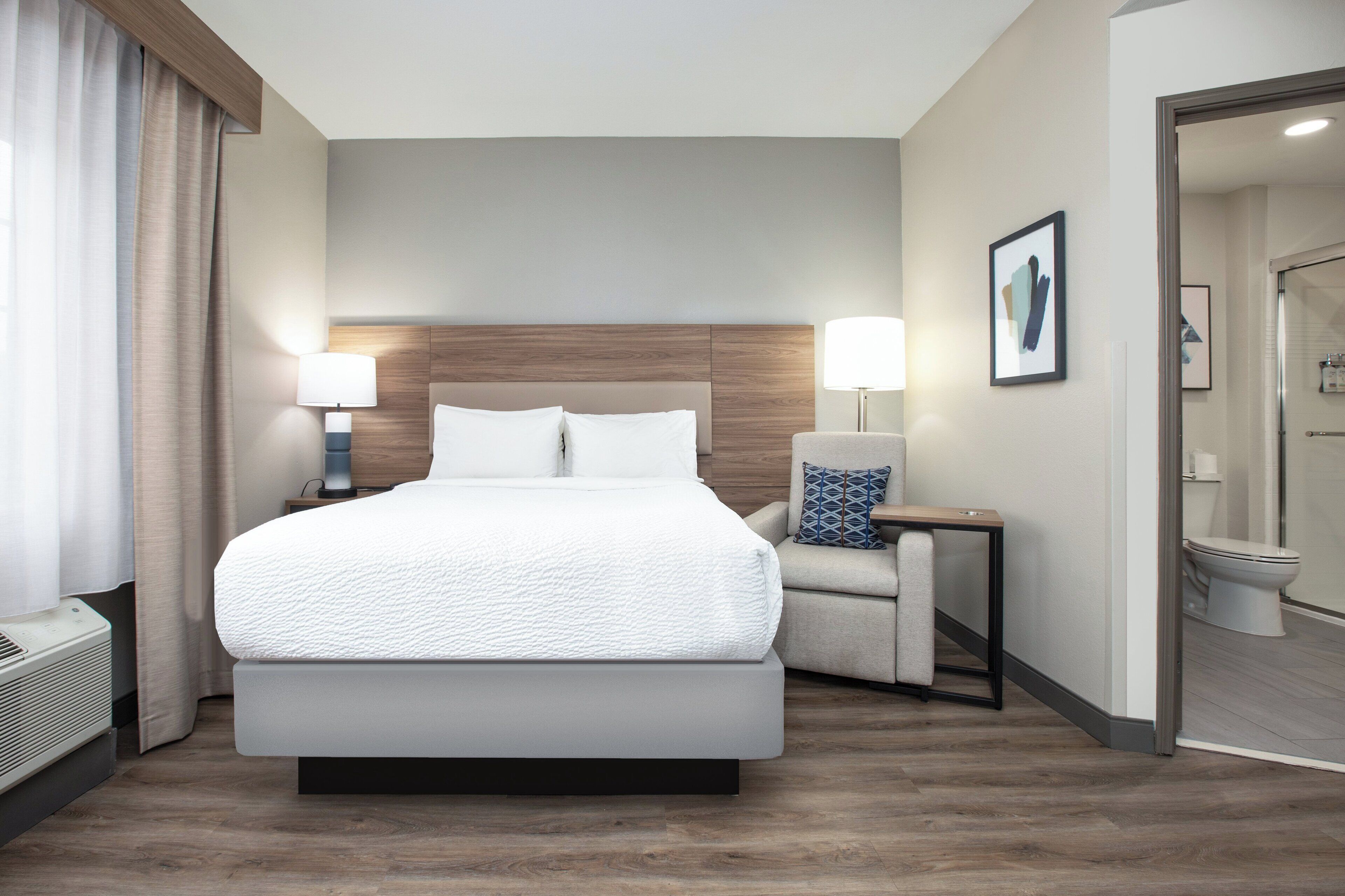 Une chambre d’hôtel moderne, dotée d’un grand lit, de lampes de chevet, d’un fauteuil et d’une salle de bain visible à travers une porte ouverte.