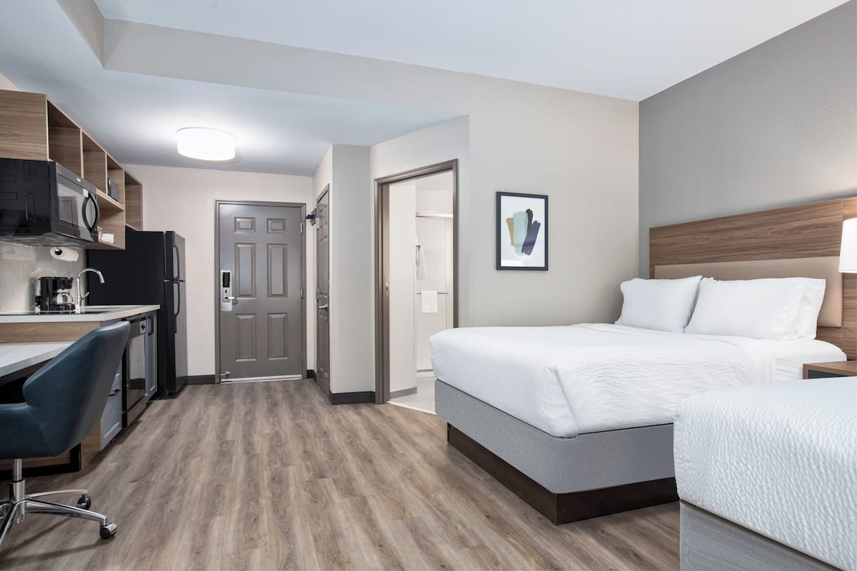 A hotel room with a bed, a desk, a chair, a microwave, a refrigerator, and a door leading to a bathroom.