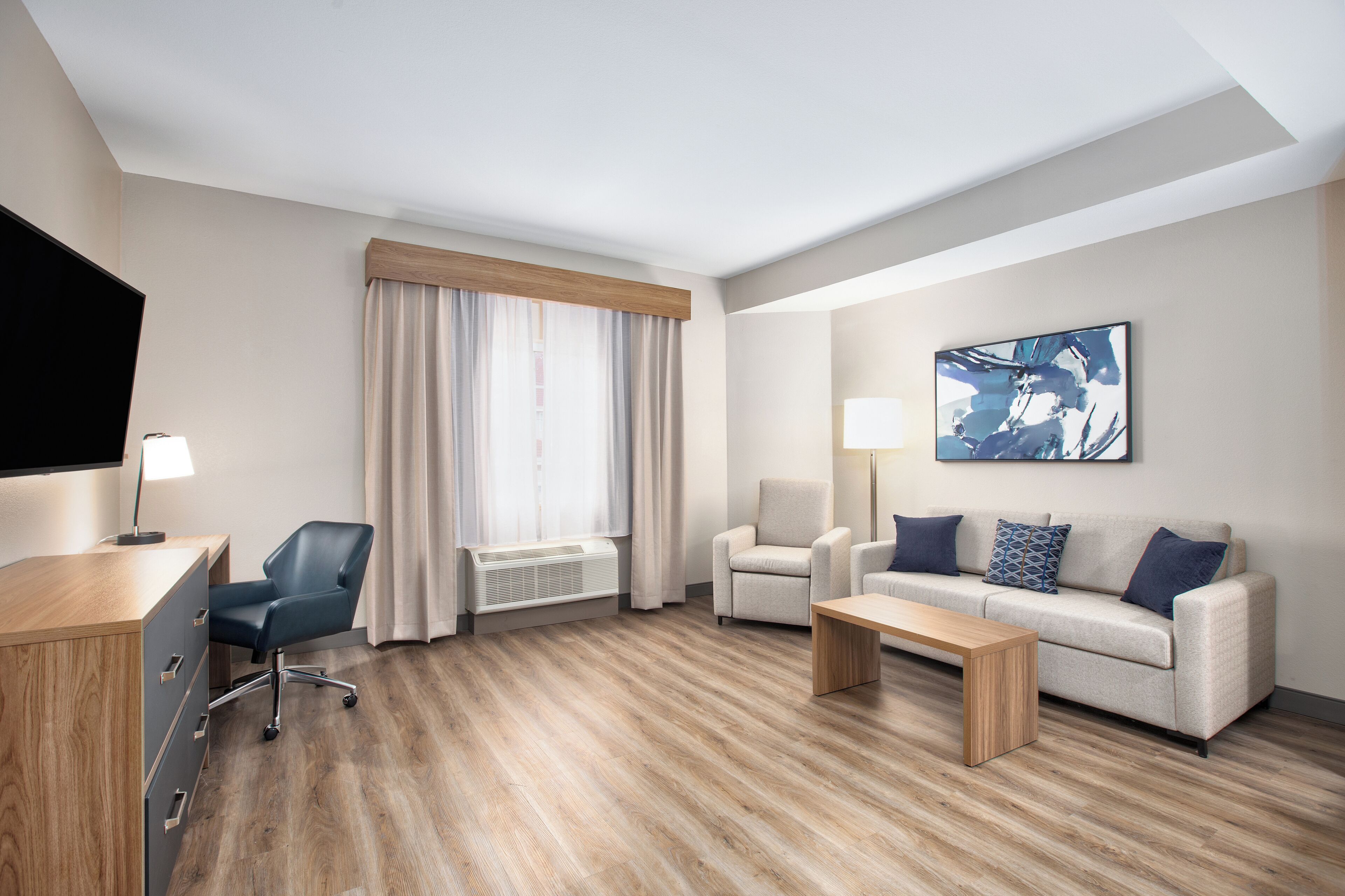 Une chambre d’hôtel avec un plancher en bois, un bureau avec une chaise, un canapé avec des coussins, une table basse, une télévision et une fenêtre avec des rideaux.