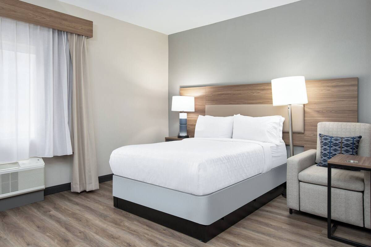 A hotel room with a large bed, two bedside lamps, a chair, and a heater.