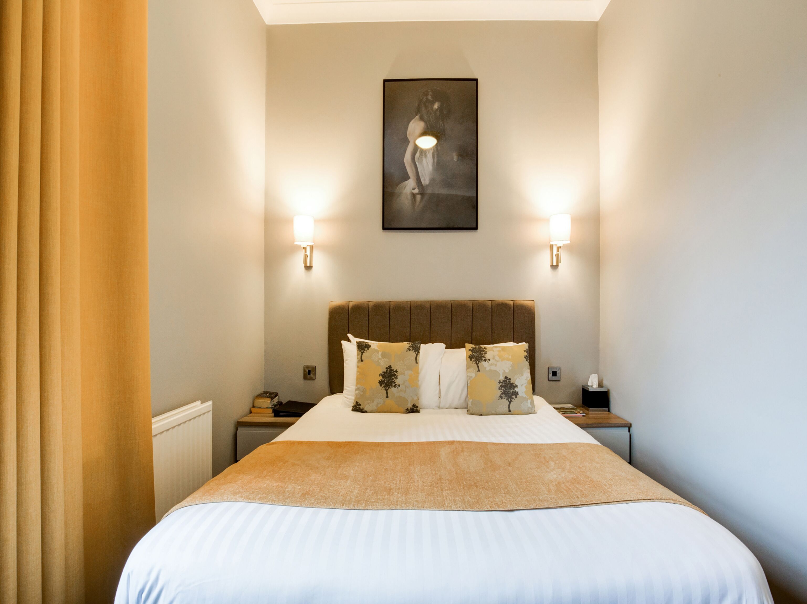 A hotel room with a large bed, two bedside tables, a headboard with decorative pillows, and wall-mounted lighting.