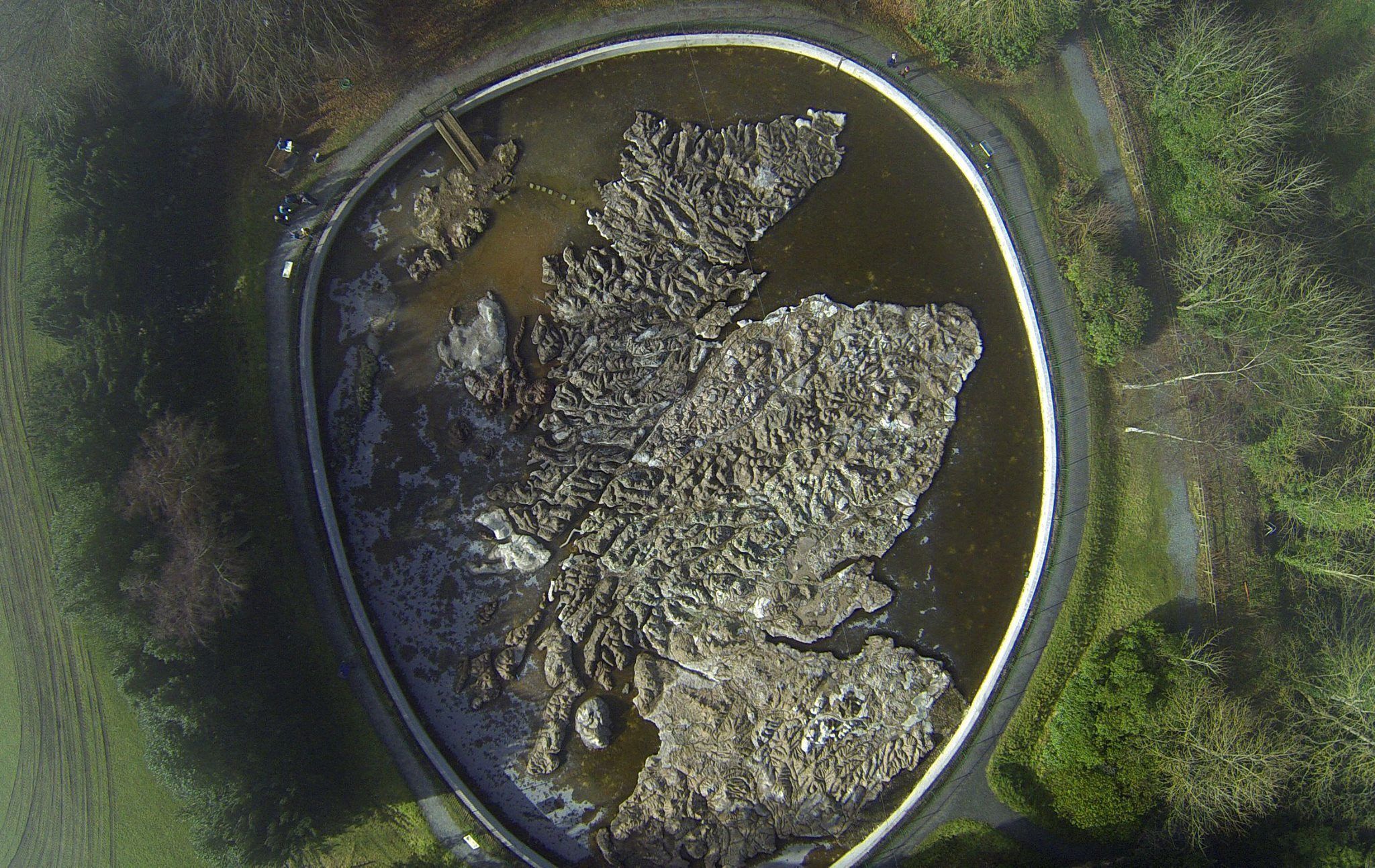 An aerial view of a circular water body with a rocky shoreline and surrounding greenery.