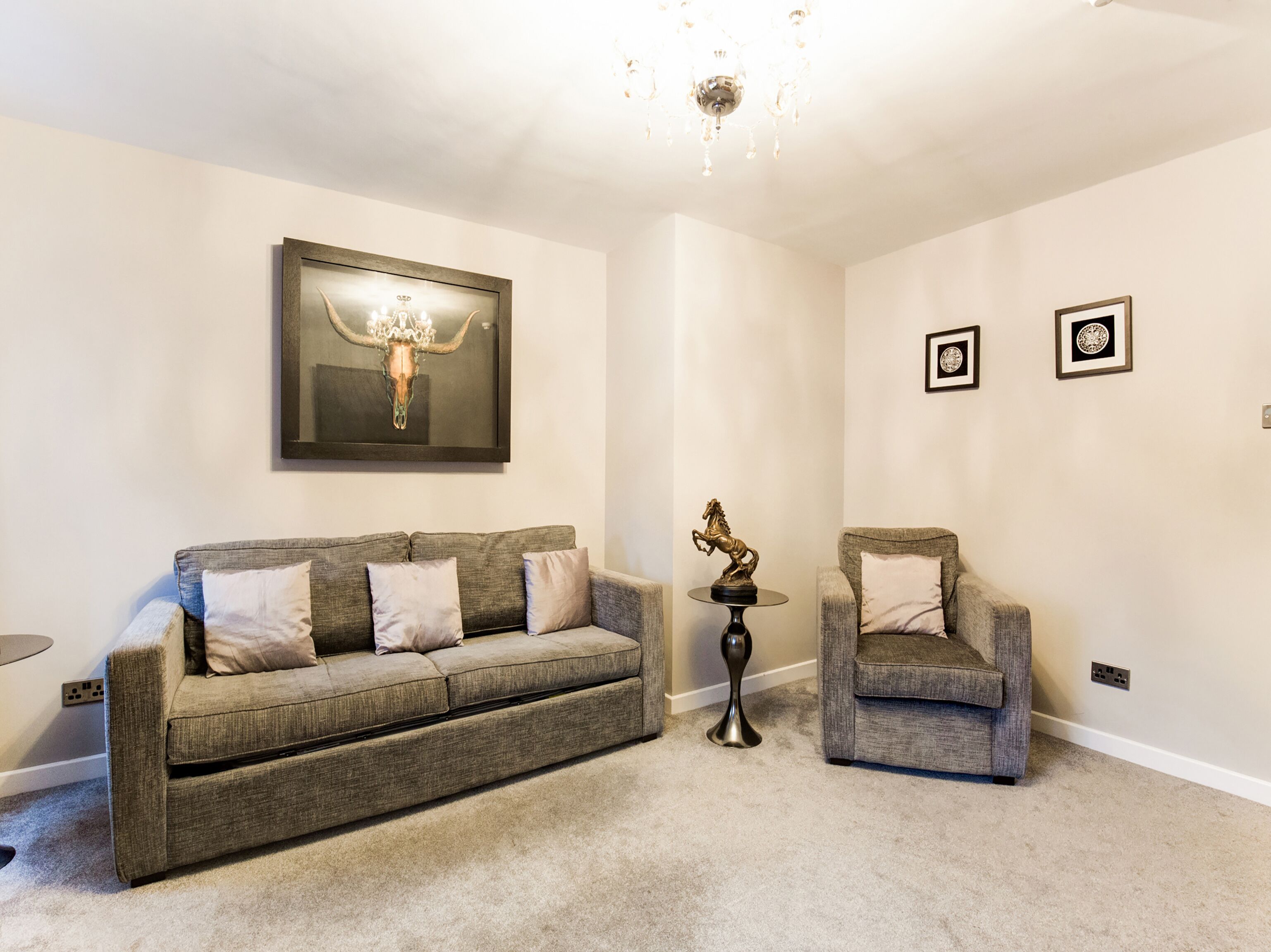 A living room with a grey sofa, a matching armchair, a small round table, and a chandelier.