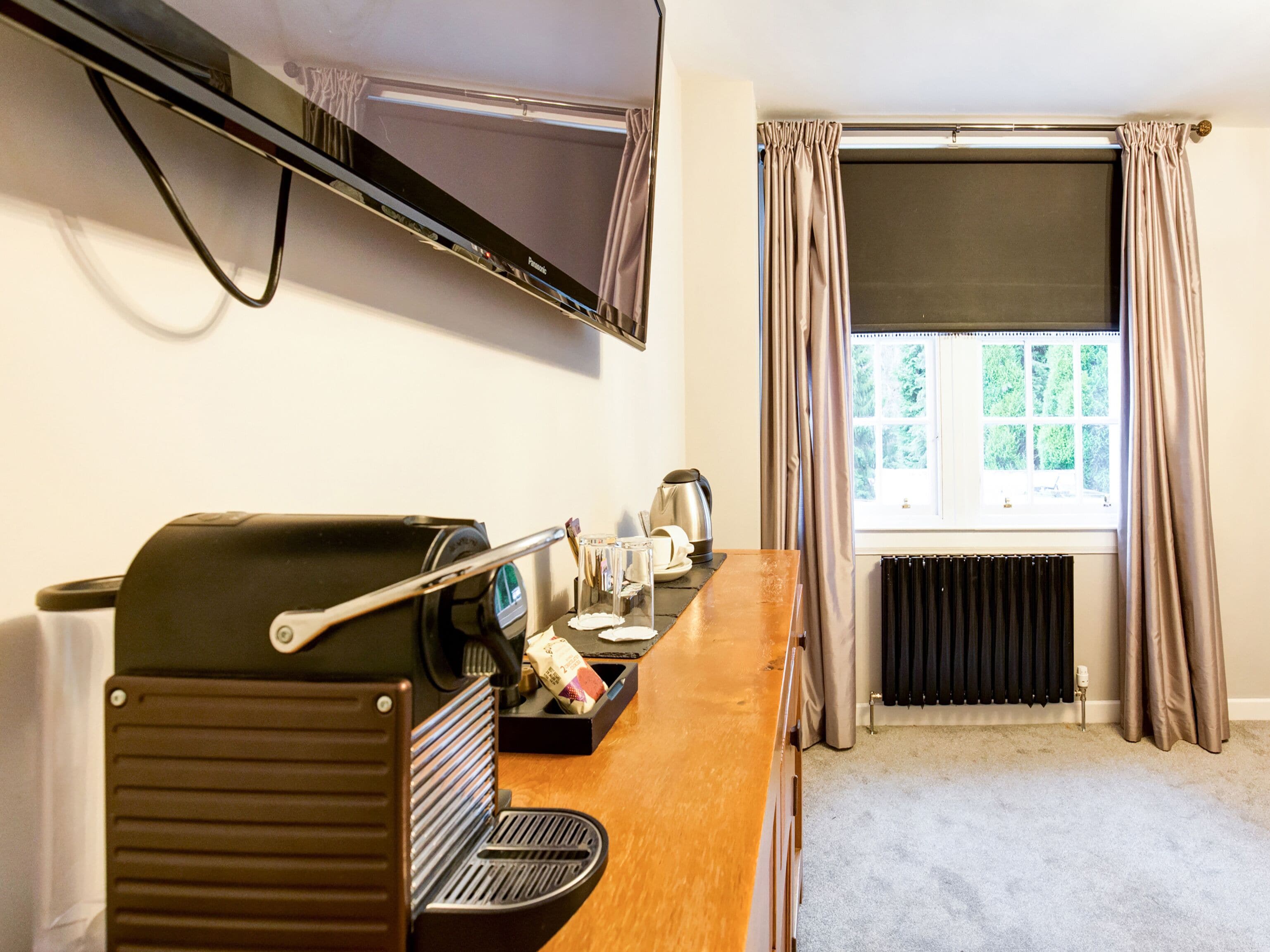 A room with a kitchenette, a flat-screen TV mounted on the wall, a window with curtains, and a radiator.