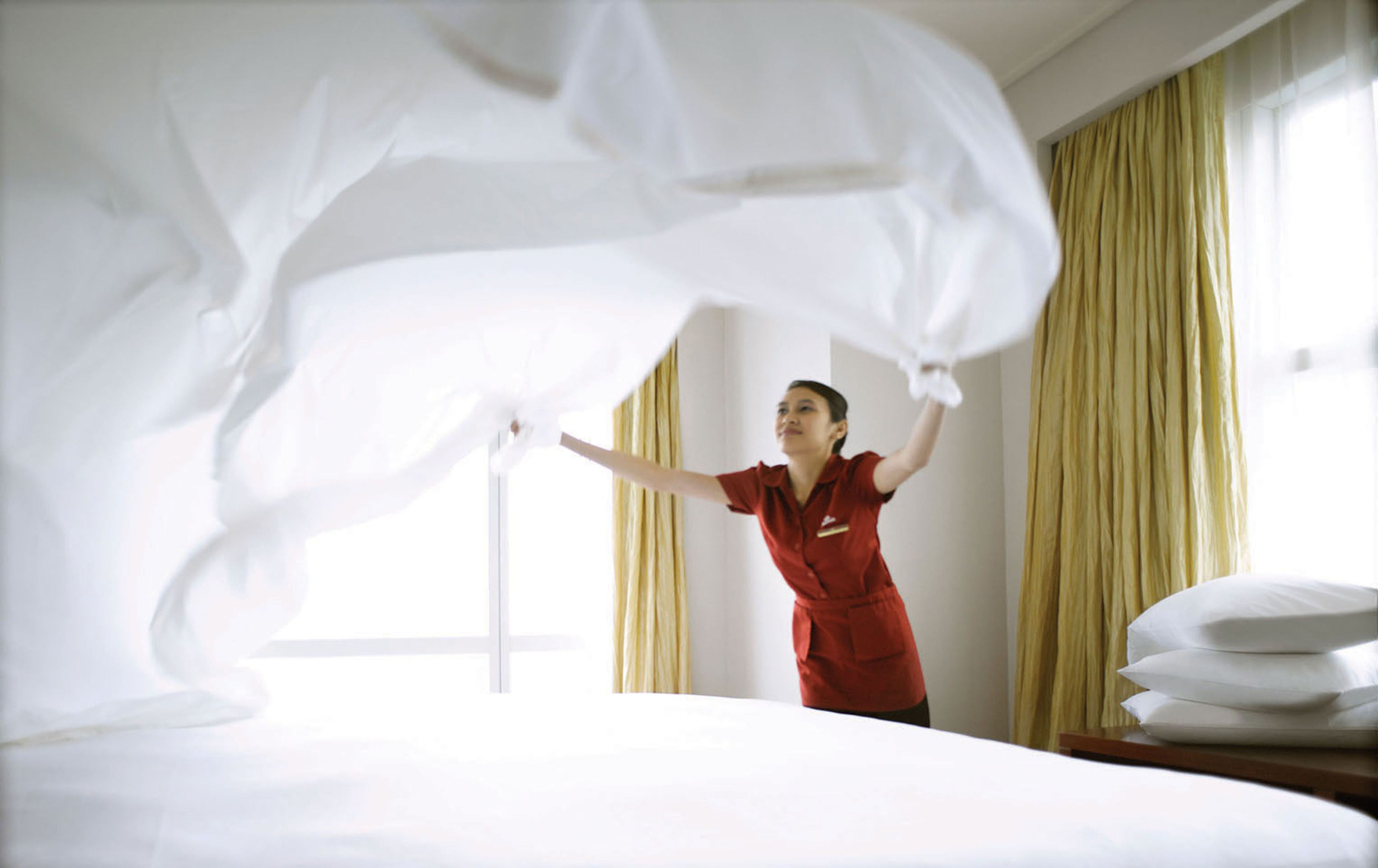 Une chambre d’hôtel avec un lit soigneusement fait, une grande fenêtre avec des rideaux et une personne vêtue d’un uniforme rouge.
