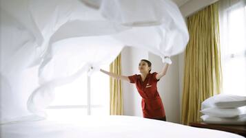 Una habitación de hotel con una cama bien hecha, un ventanal con cortinas y una persona con uniforme rojo.