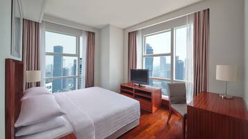 Habitación de hotel con cama, televisor sobre un atril de madera, escritorio, silla y ventanas amplias con vista a la ciudad.