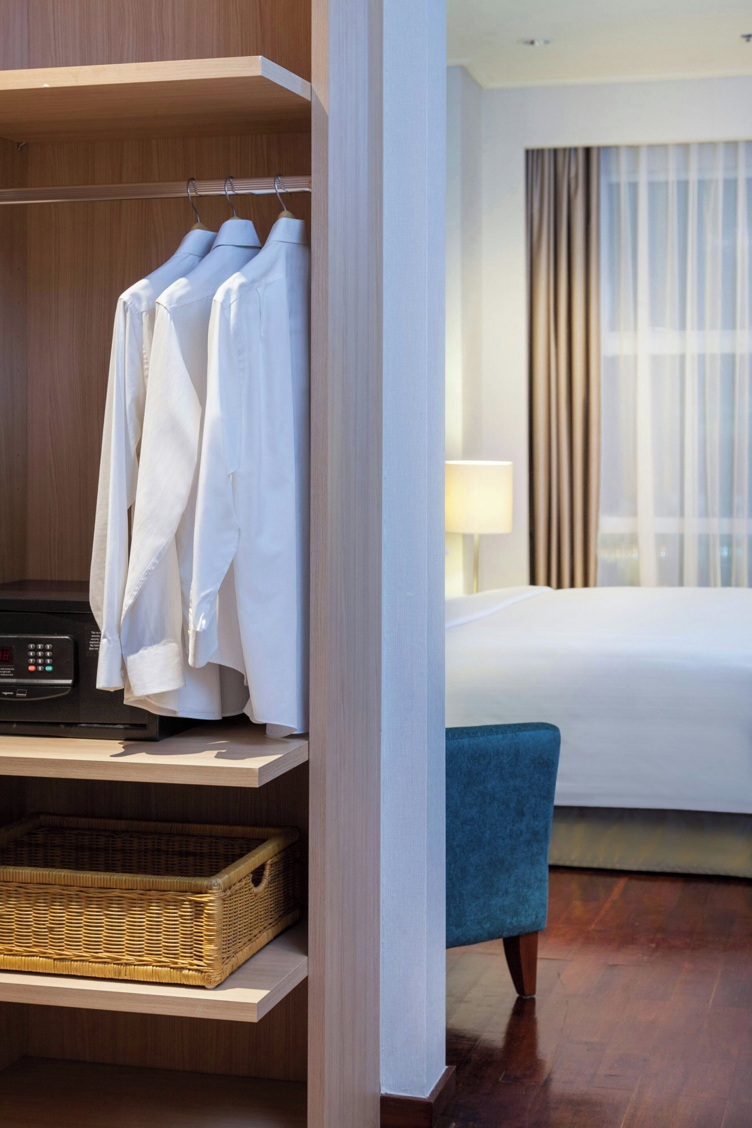 A hotel room with a bed, a blue chair, a wardrobe with hanging clothes, and a digital clock on a shelf.