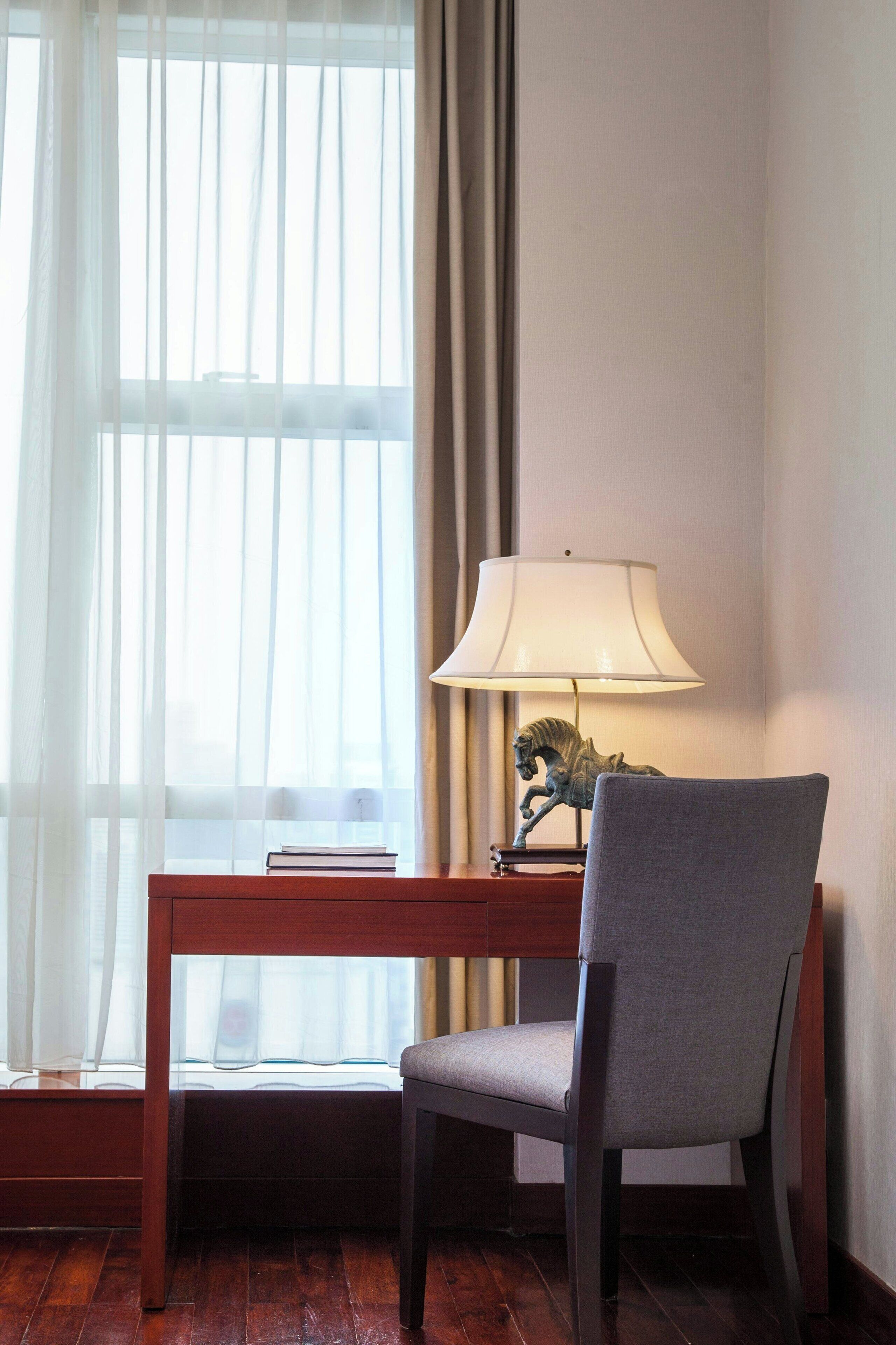 Une pièce avec un bureau en bois, une chaise grise, une lampe et une fenêtre ornée de rideaux légers.