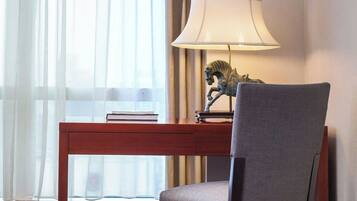 A room with a wooden desk, a grey chair, a lamp, and a window with sheer curtains.