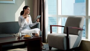 Eine Frau sitzt auf einem grauen Sofa in einem modernen Raum mit einem großen Fenster, einem Holztisch mit einem Croissant und einem Tablet.