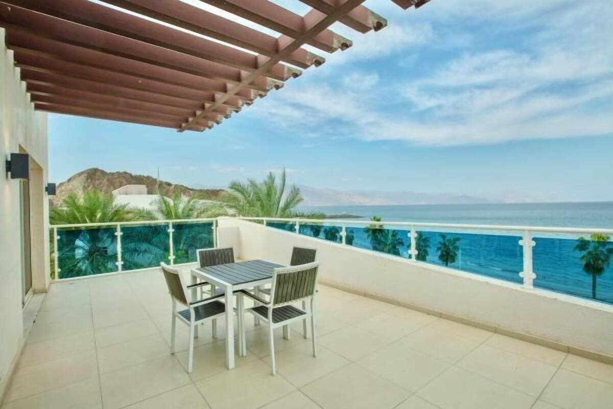 A balcony with a table and chairs, overlooking a body of water and a mountainous landscape.