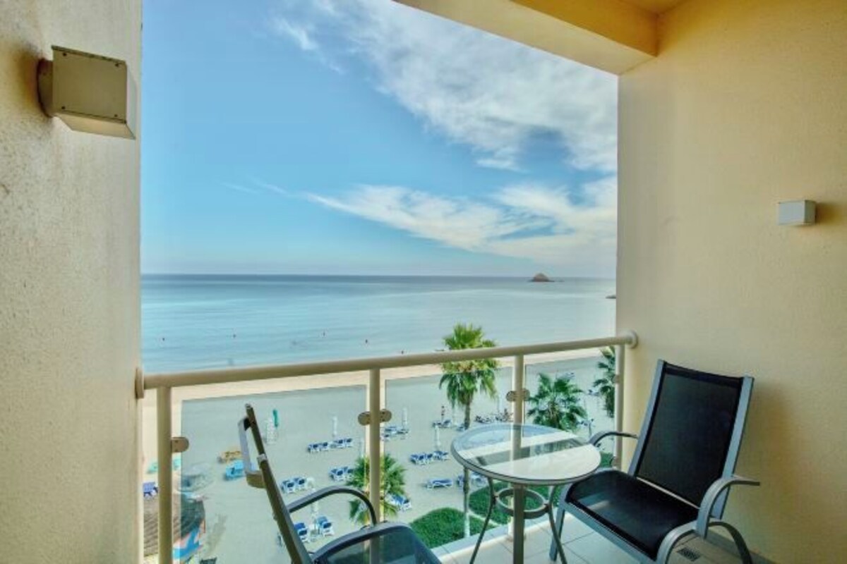 A balcony with a glass table and chairs, overlooking a beach with palm trees and parked cars.
