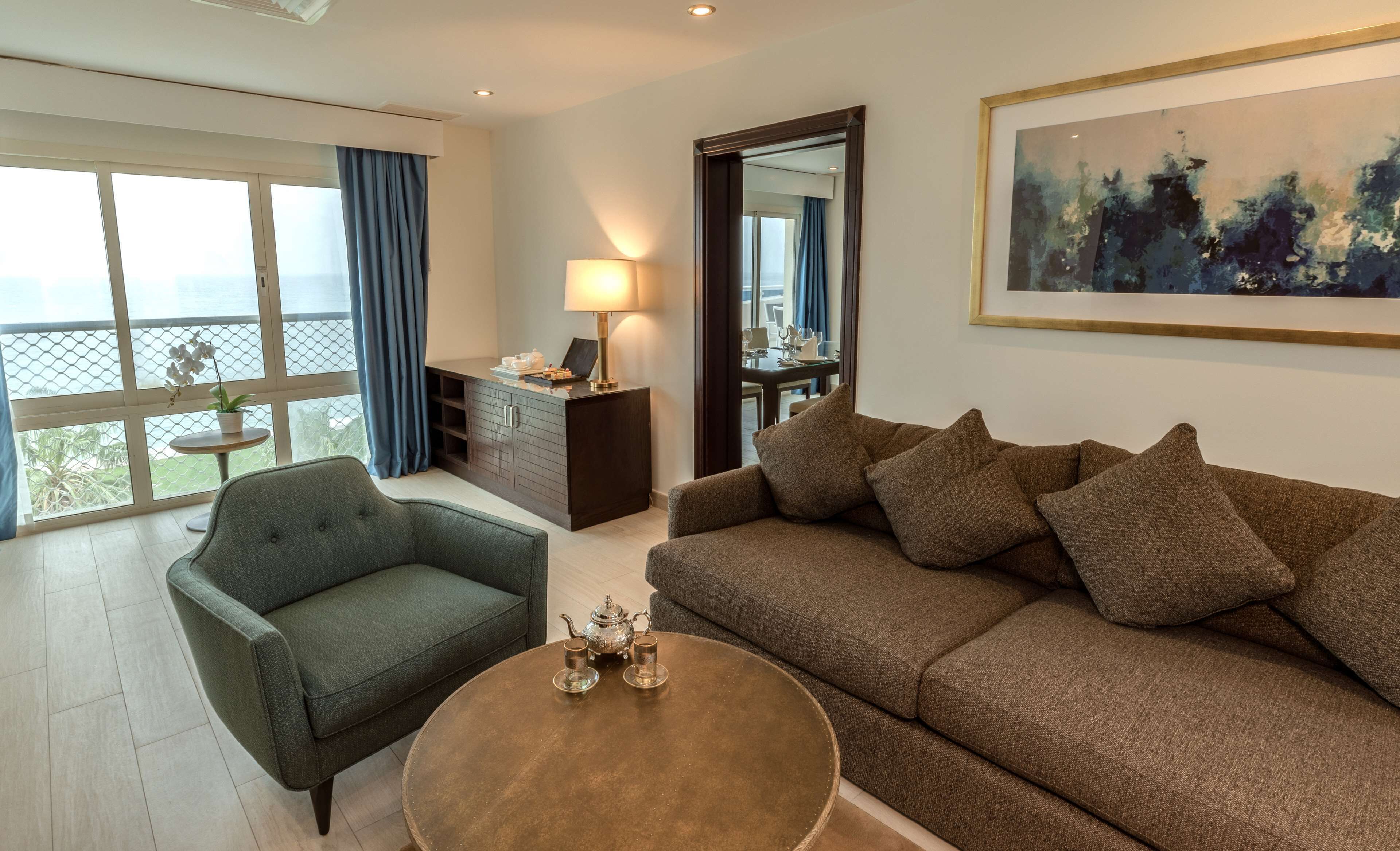 A modern living room with a sofa, armchair, and coffee table. There is a dining area with a table and chairs, and a large window offering a view.