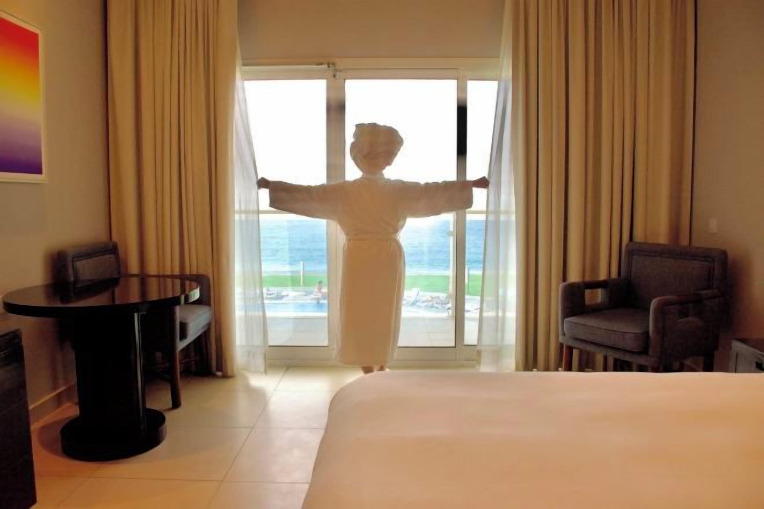 Habitación de hotel con cama, una mesa, dos sillas y vista al océano a través de un ventanal grande.