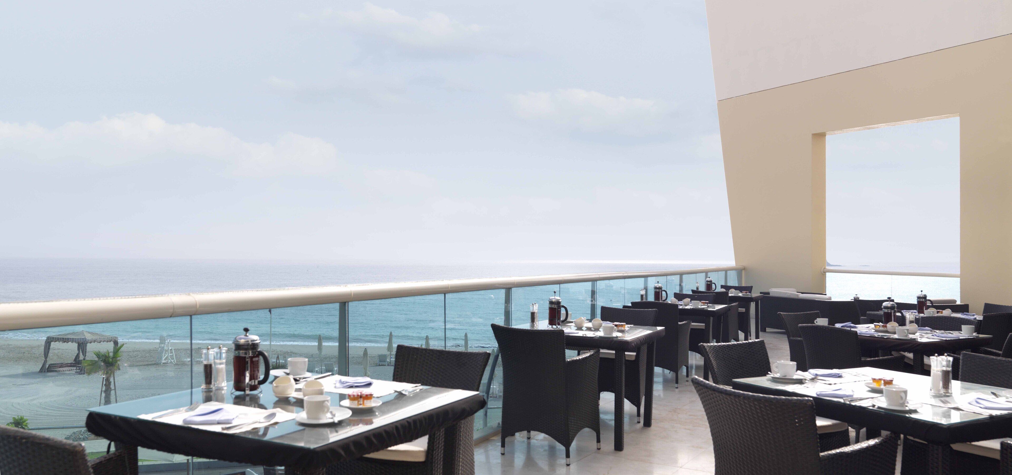 A restaurant with tables and chairs, a view of the ocean, and a balcony railing.