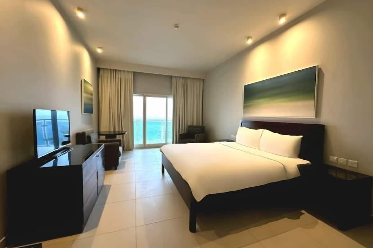 A modern hotel room with a large bed, a TV, a desk, and a view of the ocean.