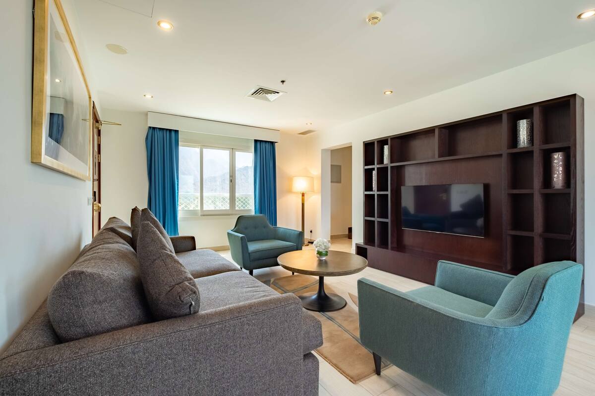 A modern living room with a grey sofa, teal armchairs, a round coffee table, and a TV on a wooden stand.
