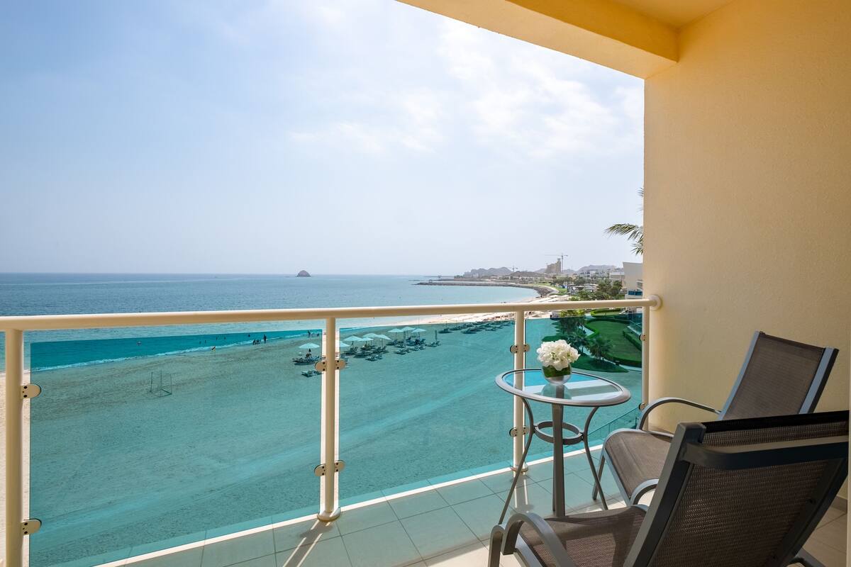 A balcony with a view of a beach and ocean, a small table with a vase, and two chairs.