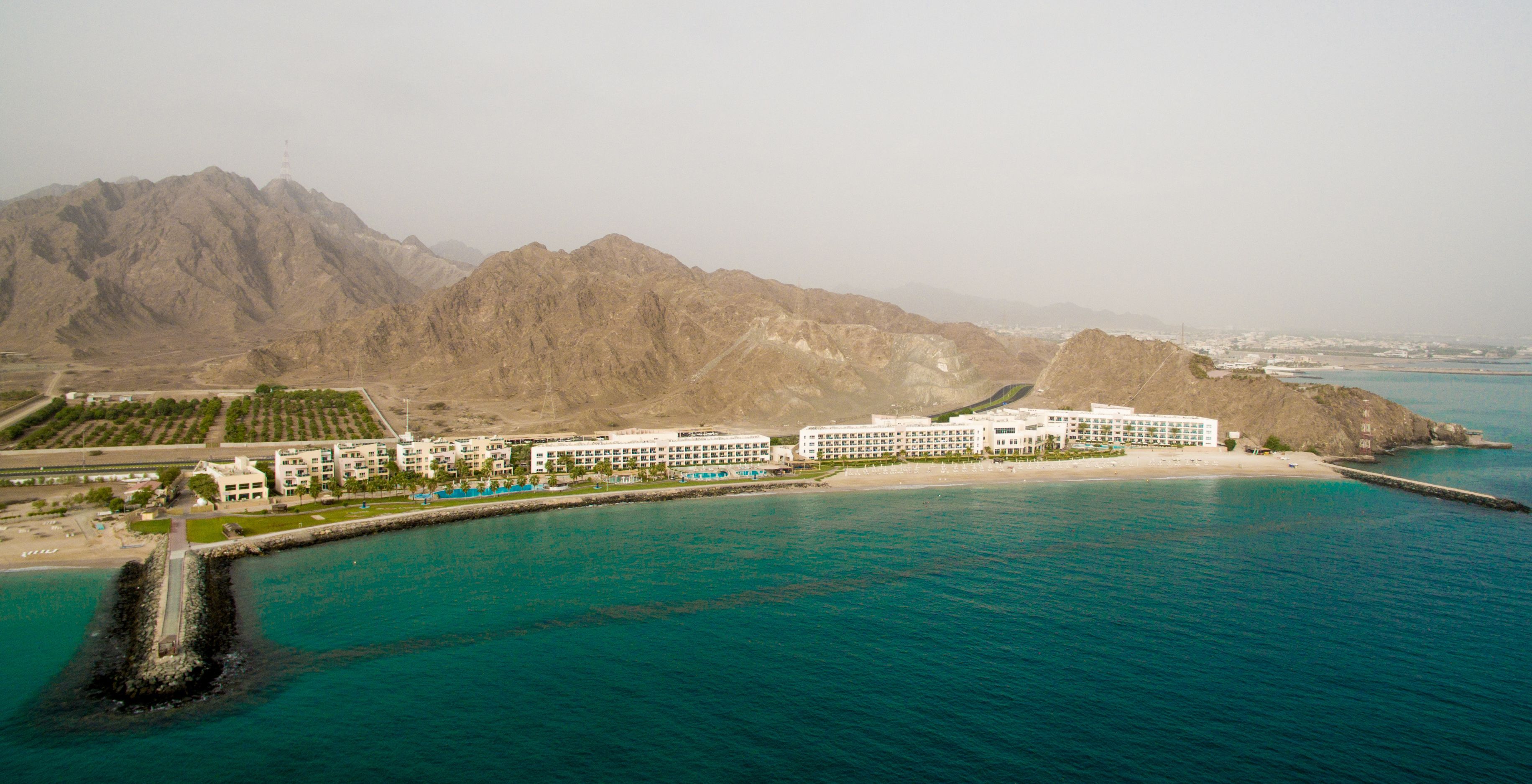 An aerial view of a coastal hotel with multiple buildings, a beach, and mountains in the background.