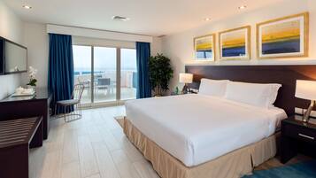 Habitación de hotel con una cama grande, un escritorio, una silla, un balcón con vistas y tres cuadros en la pared.