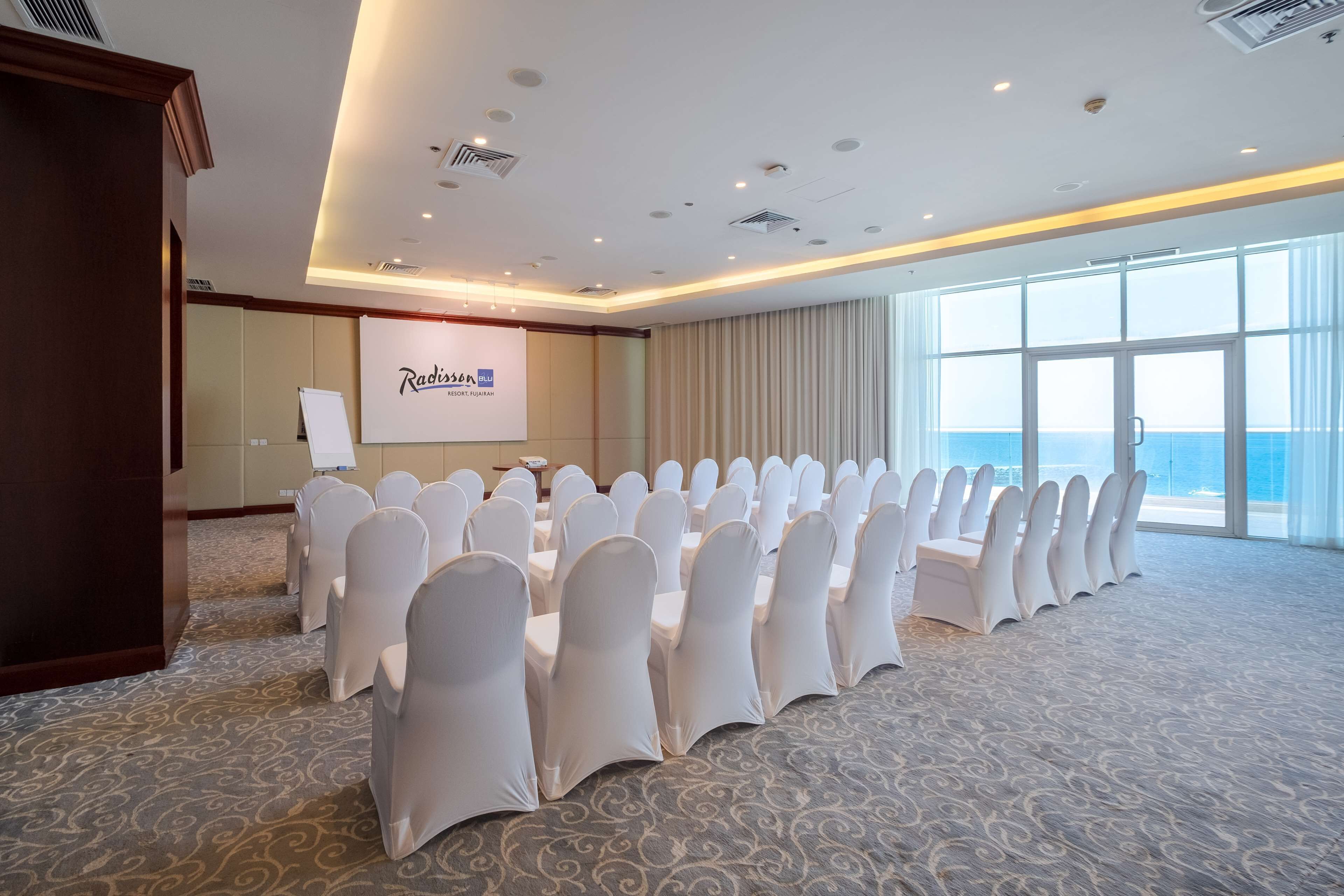 A conference room with rows of white chairs, a projection screen, and a large window with a view of the sea.