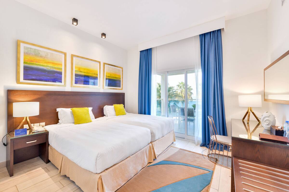 A hotel room with a large bed, a desk, a chair, and a balcony with blue curtains.