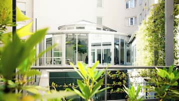 Una estructura de vidrio tipo invernadero con diseño moderno, rodeada de vegetación y contigua a un edificio con amplias ventanas.