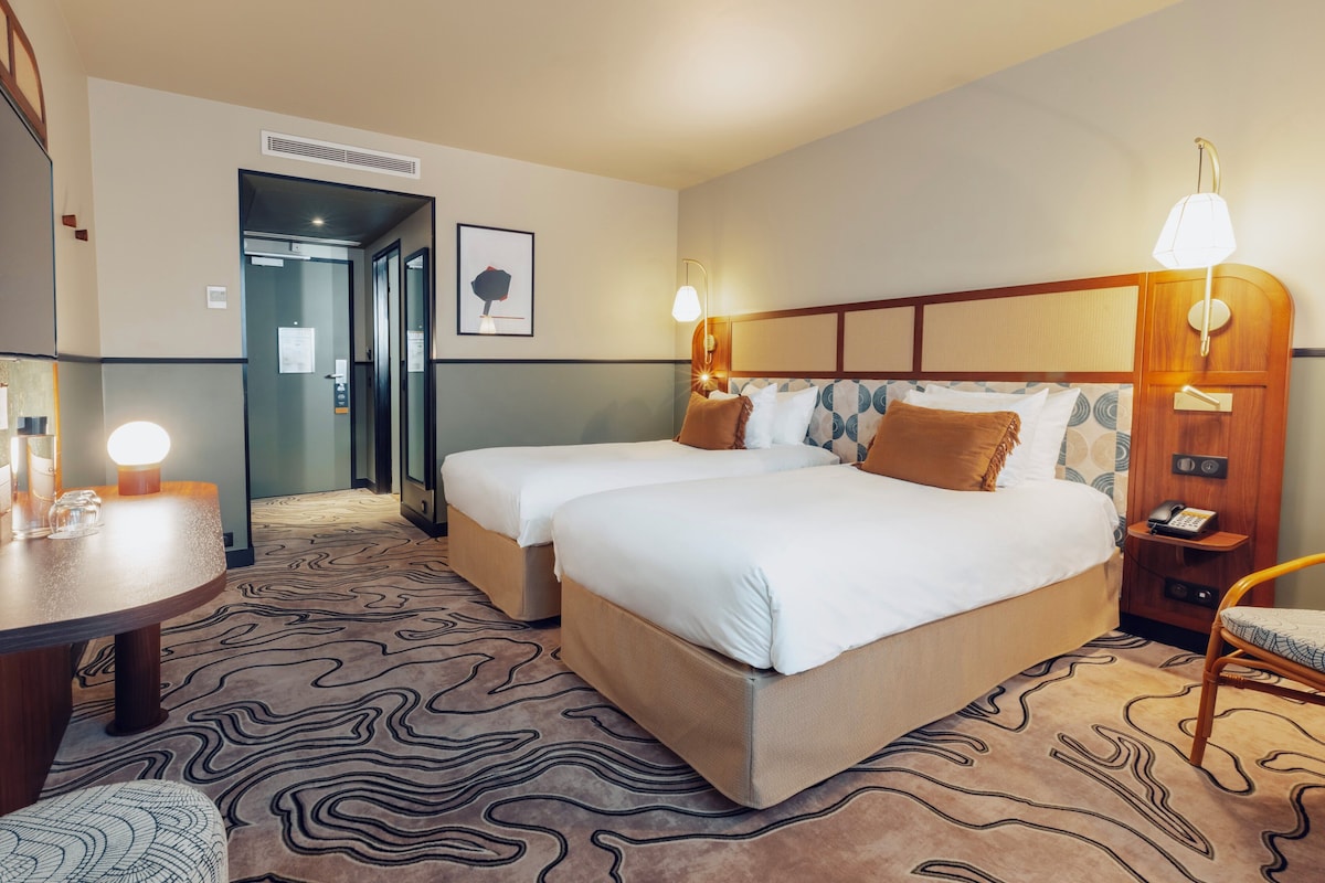A hotel room with two beds, a desk, and a chair. There is a door leading to a bathroom and a hallway.